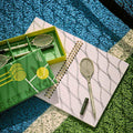 Set de bolígrafos con forma de raqueta de tenis sobre una libreta abierta, junto a su caja de presentación, en una pista de tenis. 