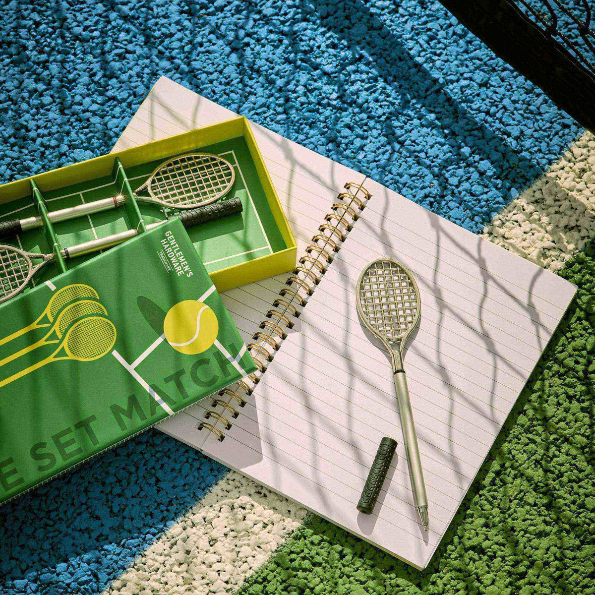 Set de bolígrafos con forma de raqueta de tenis sobre una libreta abierta, junto a su caja de presentación, en una pista de tenis. 